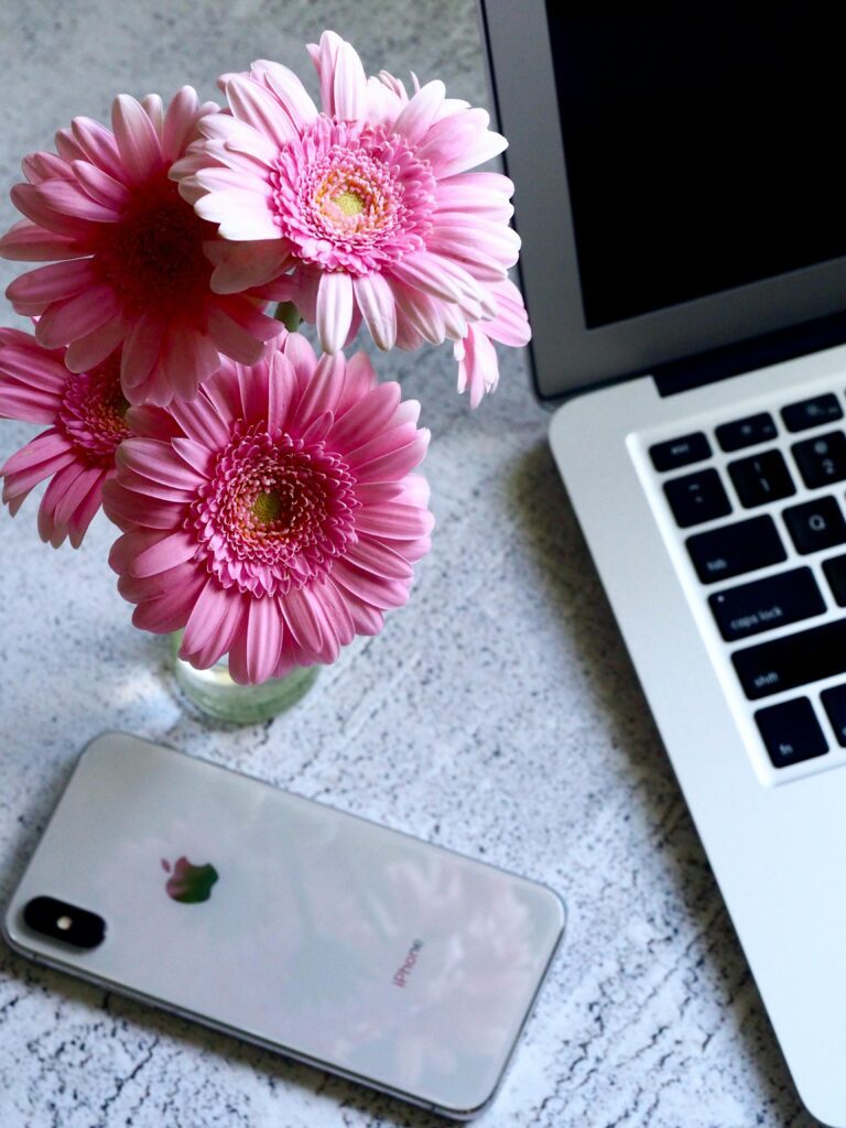 A modern workspace featuring pink gerbera flowers, a laptop, and a smartphone on a textured desk.